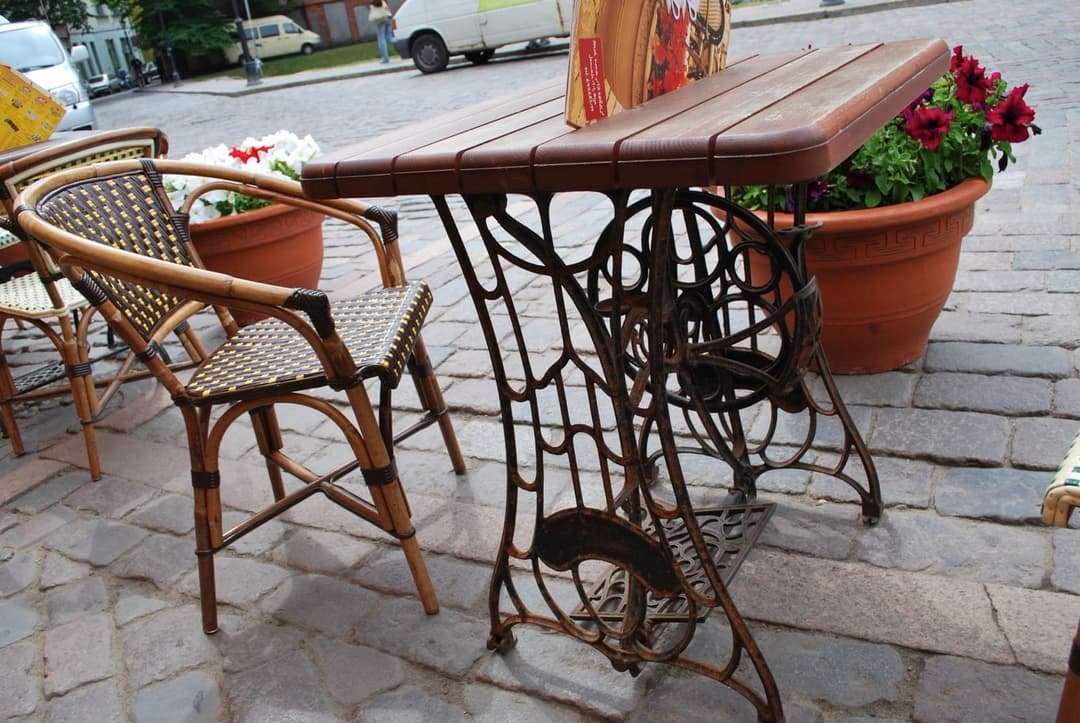 A cast-iron sewing machine treadle base with a slatted wooden top used as a table outdoors on cobblestones, beside a rattan chair, with terracotta pots of red and white petunias behind