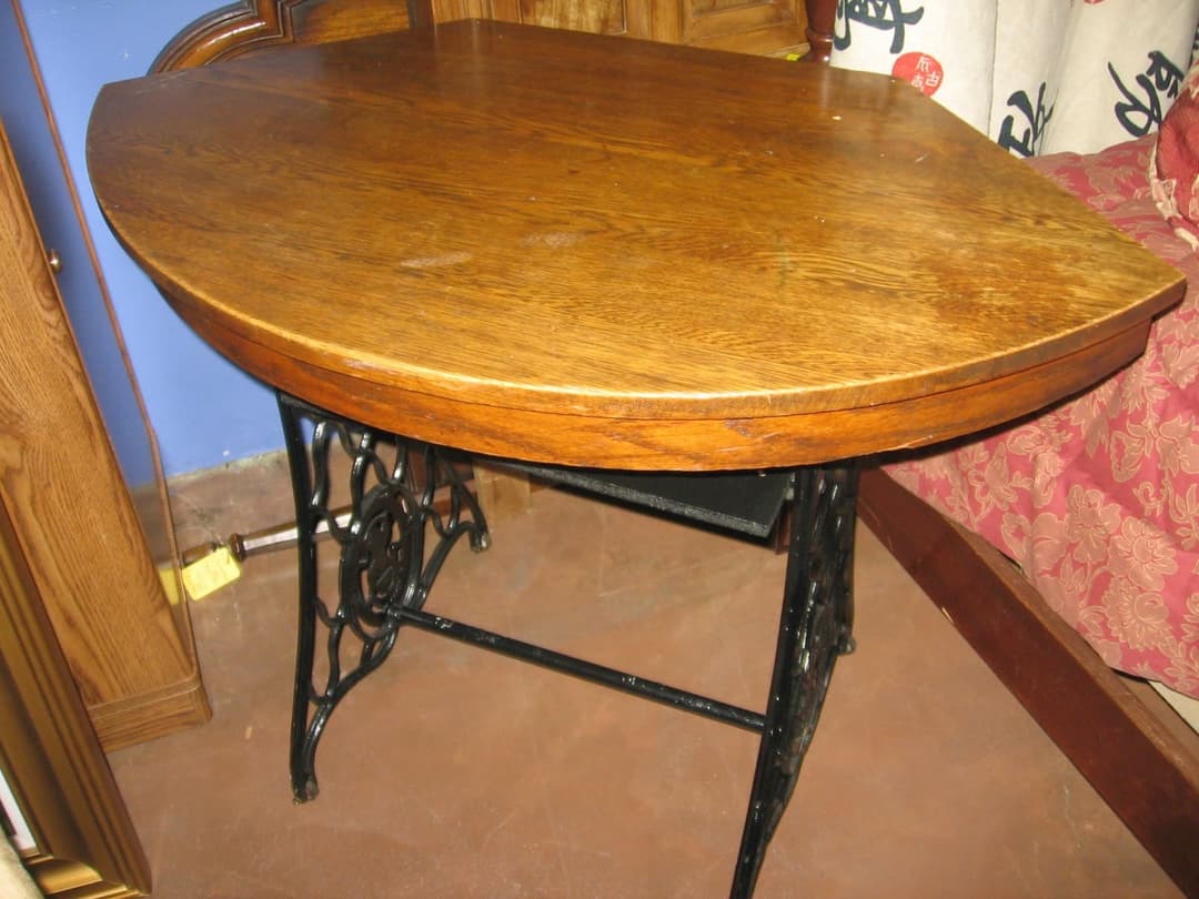A black cast-iron sewing machine treadle base with scrollwork sides, topped with a D-shaped oak board to make a small table or desk, standing in a shop