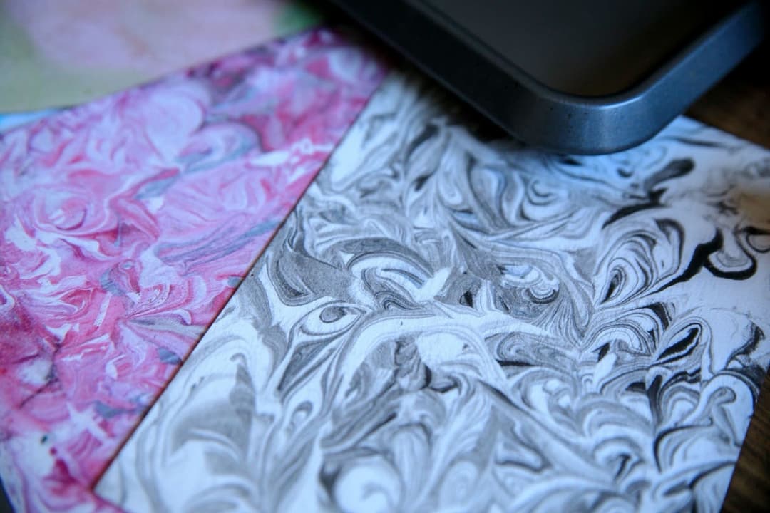 Two cards marbled with shaving cream and ink, one with grey and black swirls and one with pink swirls, on a wooden table beside a grey baking tray